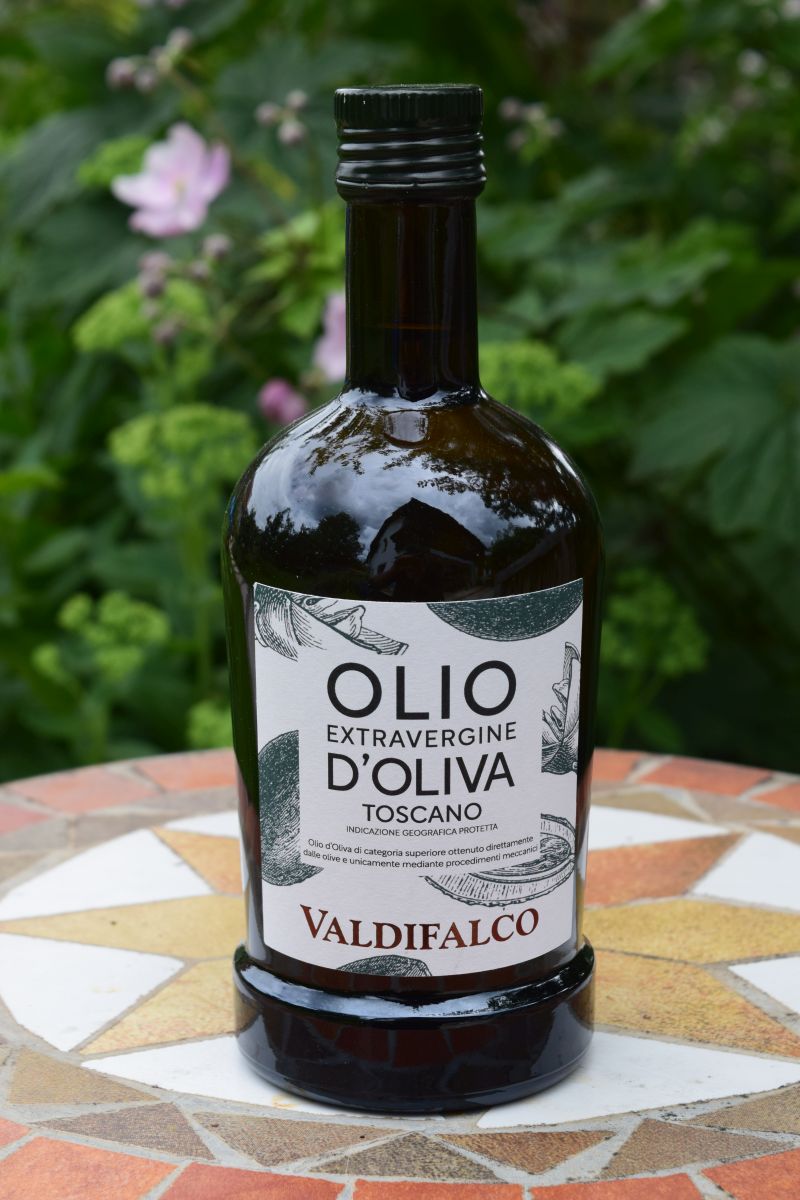 das Bild zeigt eine Flasche Olivenöl Extra Vergine im Hintergrund sieht man Pfanzen.