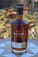Das Bild zeigt die durchsichtige kegelförmige Glasflasche mit dem braunen Rum. Die Aufschrift Walcher Südtiroler Gutsbrennerei ist zu erkennen. Im Hintergrund verschwommen Teile eines Gartens.